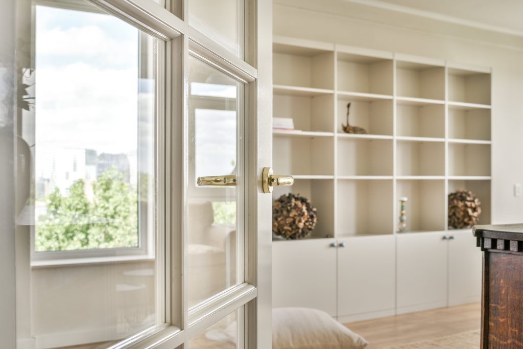 A well-lit home interior featuring an open door with a glass panel, leading to a room with built-in shelving and a window with a scenic view.