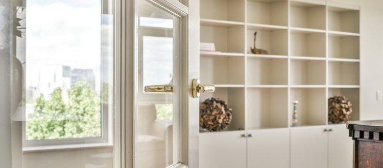 A well-lit home interior featuring an open door with a glass panel, leading to a room with built-in shelving and a window with a scenic view.